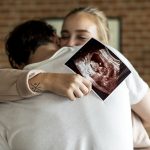 Coppia che si abbraccia emozionata, in cui la donna tiene in mano un’ecografia effettuata appena dopo i primi giorni dai sintomi della gravidanza