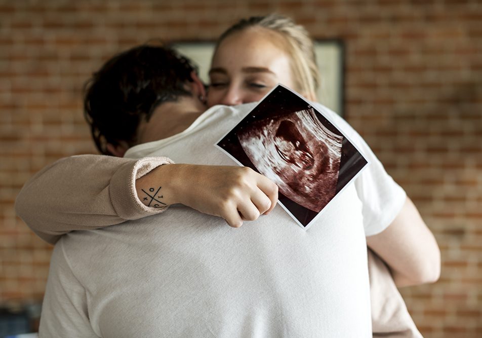 Coppia che si abbraccia emozionata, in cui la donna tiene in mano un’ecografia effettuata appena dopo i primi giorni dai sintomi della gravidanza