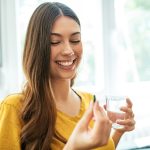 Donna sorridente che assume una compressa di penicillina, insieme a un bicchiere d’acqua, nel salotto di casa