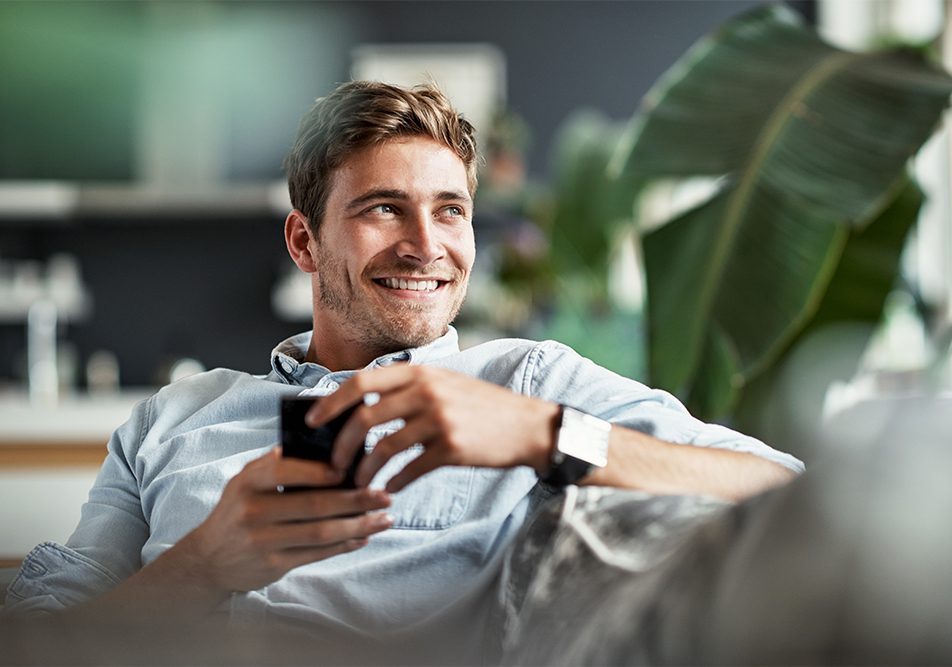 Uomo seduto rilassato sul divano di casa che prenota un check-up medico direttamente dal suo smartphone