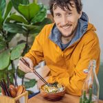 Uomo seduto ad un tavolo accanto ad una pianta, che mangia una bowl di riso e salmone, integrando l’Omega 3 e i benefici di tale nutriente attraverso un’alimentazione sana