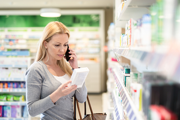 Donna in farmacia al telefono con il medico che riceve informazioni sui possibili effetti dell’allergia ai farmaci mentre legge il retro di una confezione