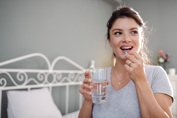 Donna sorridente ai bordi del letto, che assume un integratore per contrastare l’amenorrea insieme a un bicchiere d’acqua