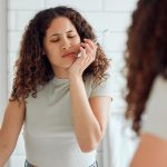 Donna davanti allo specchio del bagno, mentre lava i denti, che avverte dolori gengivali, uno dei sintomi della malocclusione dentale