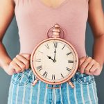 Donna con abiti trendy che tiene sul suo grembo un orologio da tavolo per segnalare il problema del ritardo del suo ciclo mestruale e interrogandosi sulle possibili cause