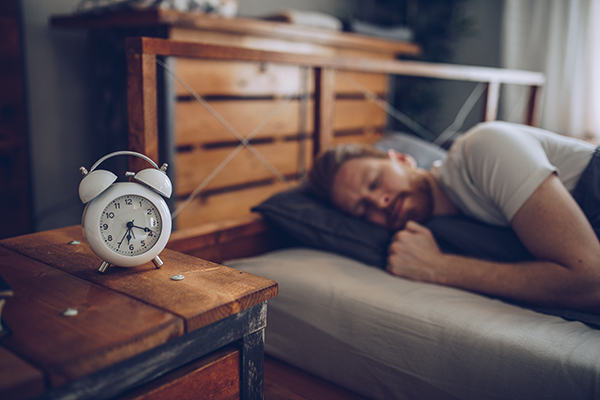 Uomo a letto al mattino, con una sveglia analogica sul comodino, che previene l’ansia cronica grazie ad un sonno regolare