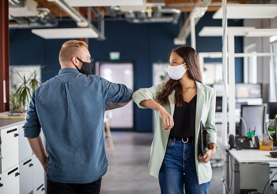 Due colleghi nel corridoio dell’ufficio che si salutano con il gomito, adottando misure di prevenzione per evitare il contagio da Covid-19