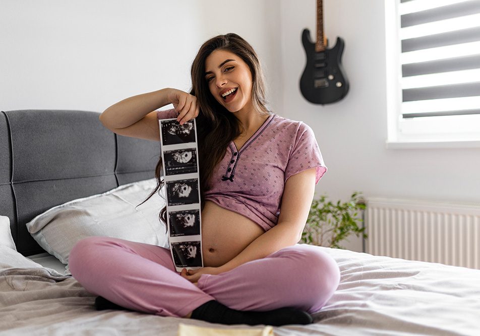 Donna incinta in pigiama che mostra felicemente le prime ecografie del nascituro, dopo avere superato i sintomi della gravidanza dei primi giorni