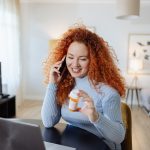 Donna seduta al tavolo del soggiorno, che domanda per via telefonica al suo medico degli esami clinici mentre tiene in mano un tubetto di medicinali