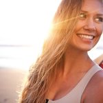 Ragazza dalla chioma bionda che surfa in spiaggia al tramonto, dopo avere applicato una protezione dei capelli dal sole