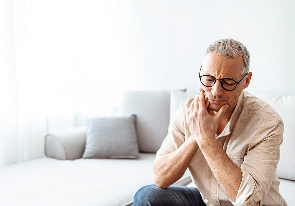 Uomo maturo seduto sul divano di casa che manifesta dolore ai denti e si tocca la mandibola infiammata