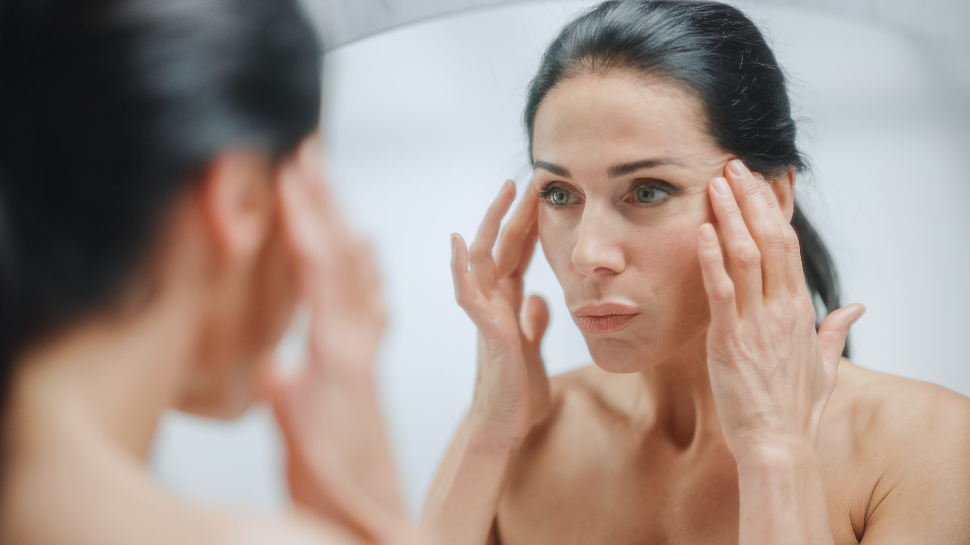 donna con capelli lunghi scuri osserva pelle viso allo specchio