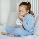 bambina con pigiama celeste seduta sul letto che tossisce