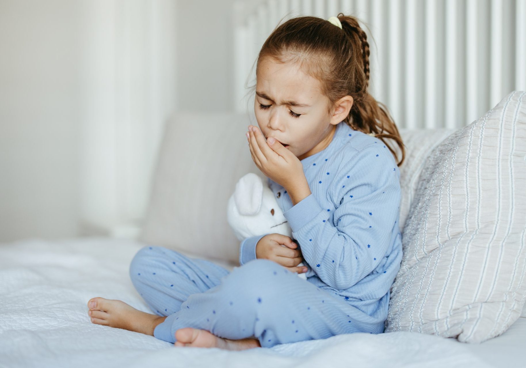 bambina con pigiama celeste seduta sul letto che tossisce